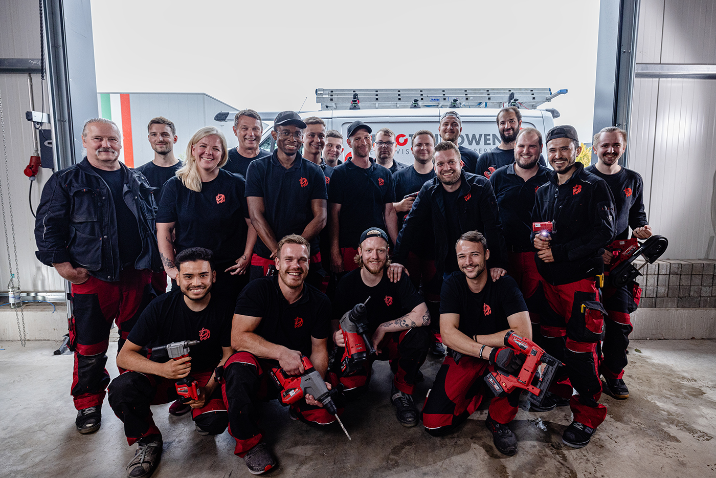 Teamfoto der Roten Löwen GmbH: Etwa 20 Mitarbeiter in schwarzen T-Shirts mit rotem Logo und roten Arbeitshosen posieren in einer Werkstatt vor einem Lieferwagen.