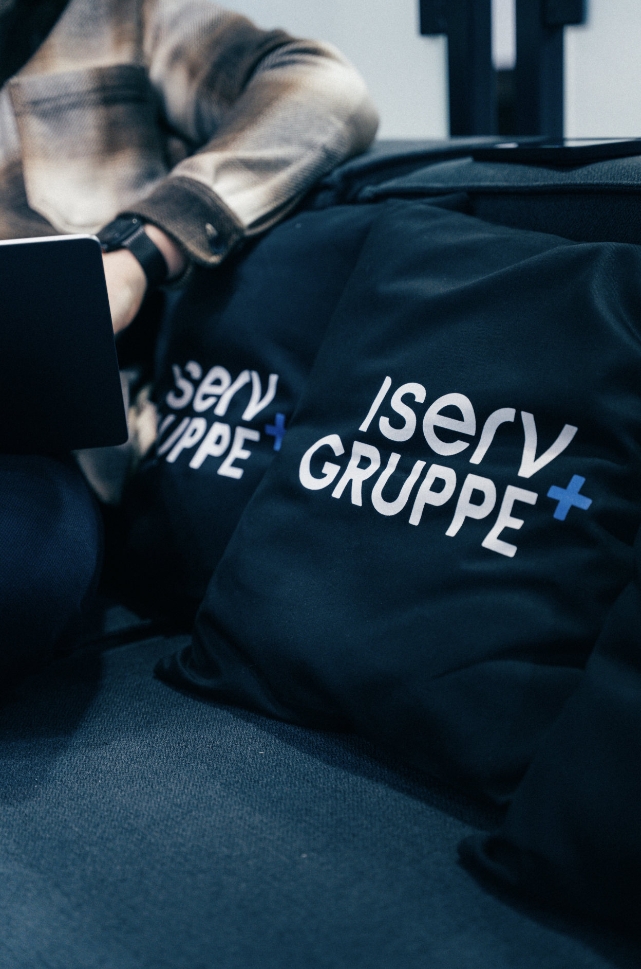 IServ Gruppe Merchandise, dunkelblaue Jacke mit weißem Logo und blauem Plus-Symbol, getragen von Person in grauer Jacke, Nahaufnahme der Markenentwicklung
