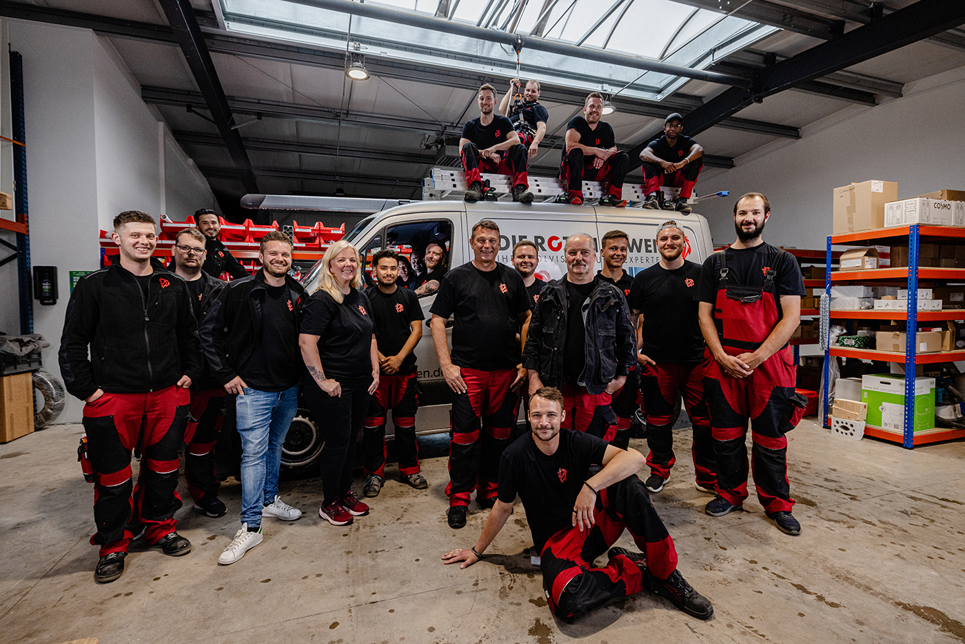 Team der Roten Löwen GmbH in schwarzen und roten Arbeitsanzügen posiert vor einem Firmenfahrzeug in einer Lagerhalle mit Regalen und Materialien.
