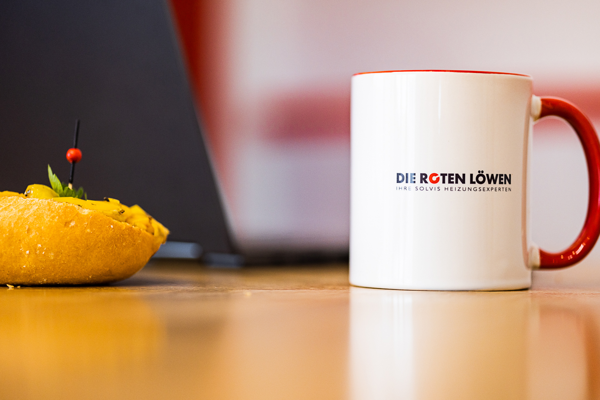 Weiße Tasse mit rotem Henkel und Logo "Die Roten Löwen, Ihre Solvis Heizungsexperten" neben einer gelben Zitrone mit Minze und roter Beere auf Holztisch