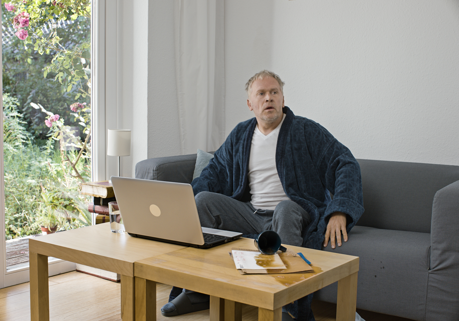 Mann sitzt auf Sofa mit Laptop vor Holztisch, helles Wohnzimmer mit Fenster und Garten, IServ Kampagne in45Minuten