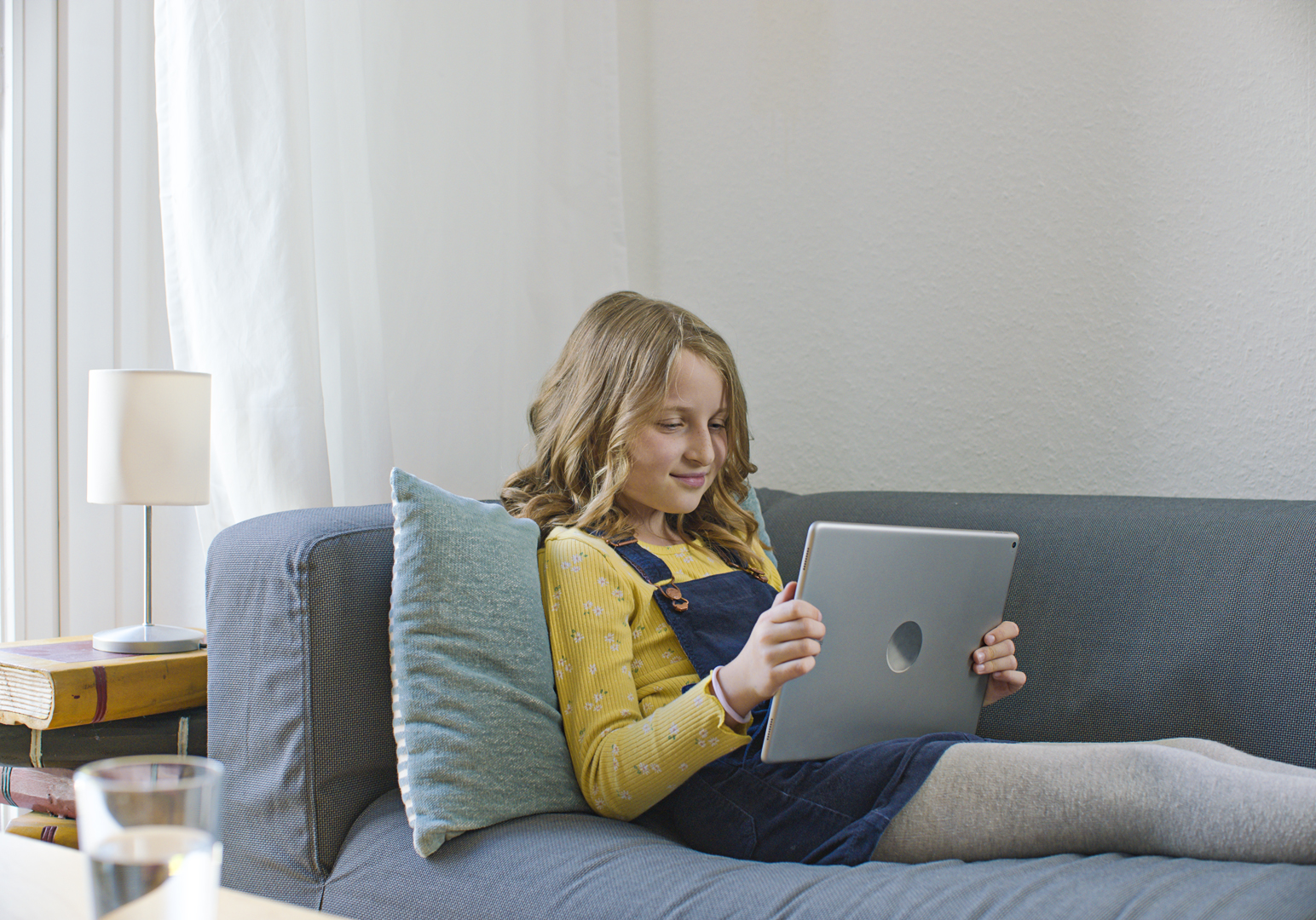 Mädchen sitzt auf Sofa und nutzt Tablet für Online-Lernen, helles Wohnzimmer mit Lampe und Büchern, IServ Kampagne in45Minuten